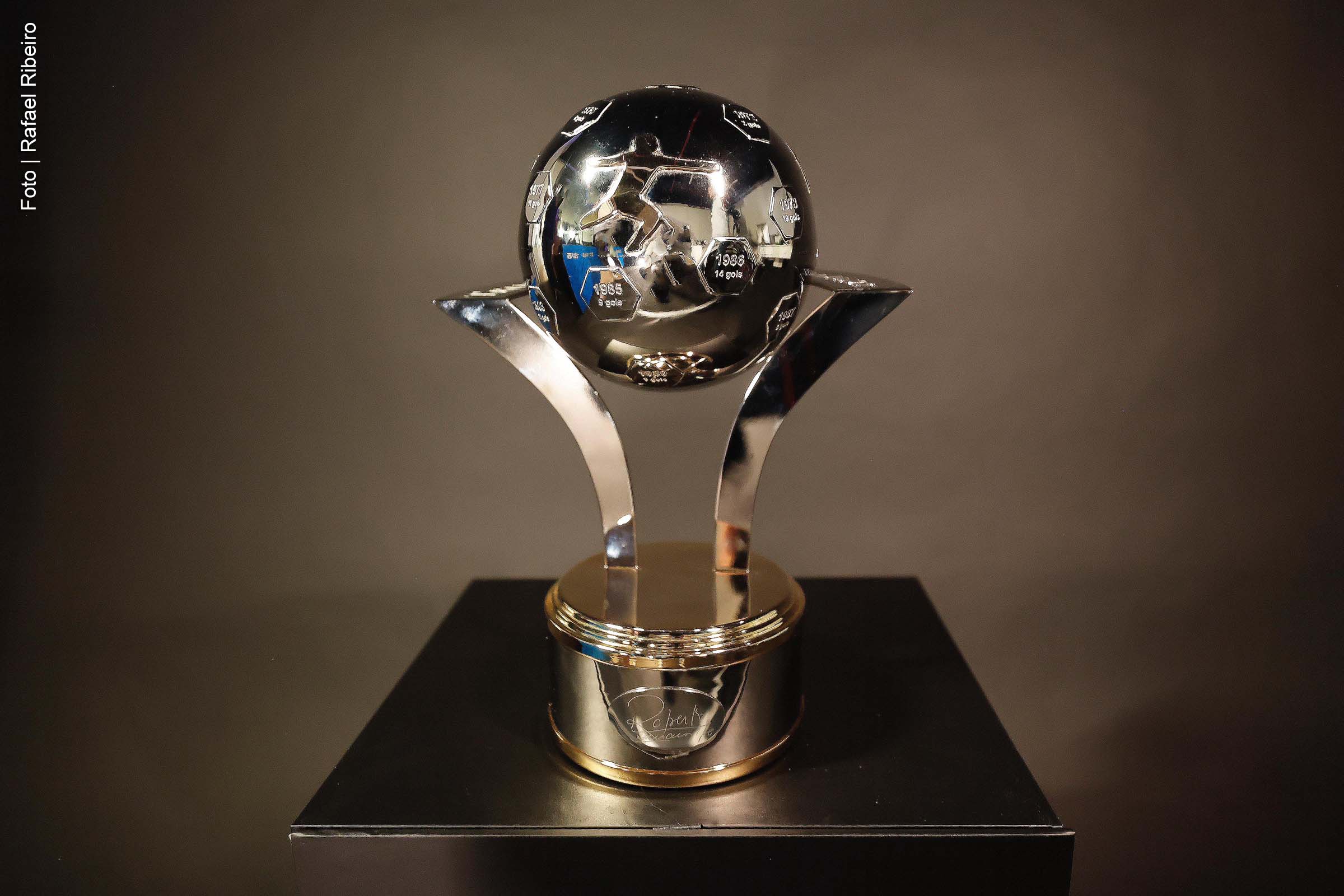 CBF apresenta troféu para artilheiro do Brasileirão, com design inspirado nos gols de Roberto Dinamite, maior goleador da competição. O prêmio reconhece a história do ex-jogador do Vasco e da Seleção Brasileira e inclui premiação em dinheiro aos vencedores. Legenda da foto: Troféu Roberto Dinamite, criado pela CBF para homenagear o maior artilheiro da história do Campeonato Brasileiro. Créditos: Rafael Ribeiro/CBF. Corpo da matéria A Confederação Brasileira de Futebol (CBF) apresentou oficialmente o Troféu Roberto Dinamite, criado para premiar os artilheiros do Campeonato Brasileiro a partir de 2024. A peça homenageia o ex-jogador Roberto Dinamite, que marcou 190 gols na história da competição, sendo até hoje o maior goleador do torneio. O troféu tem design único, com gomos representando os anos em que o jogador atuou no Brasileirão, detalhando o número de gols marcados em cada temporada. A peça ainda exibe o autógrafo do ex-atleta, reconhecido por seu desempenho nos anos 1970 e 1980, tanto no Vasco quanto na Seleção Brasileira. Durante a cerimônia de lançamento, o presidente da CBF, Ednaldo Rodrigues, destacou o significado do prêmio. "Essa é uma homenagem especial ao maior artilheiro do Campeonato Brasileiro. Roberto Dinamite merece ser eternizado, e por isso o troféu será entregue ao artilheiro de cada edição a partir de agora", afirmou. A disputa pela artilharia no Brasileirão Betano 2024 está acirrada. No momento, Alerrandro, do Vitória, e Yuri Alberto, do Corinthians, lideram com 14 gols. Estevão, do Palmeiras, aparece logo atrás, com 13. Outros jogadores, como Wesley (Internacional), Luciano (São Paulo) e Raphael Veiga (Palmeiras), seguem na disputa, com 11 gols cada. Pedro, do Flamengo, também tem 11 gols, mas está fora da competição devido a uma lesão que exigiu cirurgia. Além do troféu, o artilheiro receberá uma premiação em dinheiro, incluindo um valor fixo de R$ 100 mil e R$ 5 mil por gol marcado. No caso de empate, todos os artilheiros receberão o prêmio. Caso a decisão ocorra no final da 38ª rodada, a cerimônia de entrega está prevista para a terça-feira (10), na sede da CBF, no Rio de Janeiro. Resumo A Confederação Brasileira de Futebol apresentou o Troféu Roberto Dinamite, uma homenagem ao maior artilheiro da história do Brasileirão. A peça, com design que remete aos 190 gols de Dinamite, será entregue ao artilheiro do campeonato, que também receberá prêmios em dinheiro. A disputa pela artilharia do Brasileirão 2024 segue acirrada, com Alerrandro, do Vitória, e Yuri Alberto, do Corinthians, empatados na liderança. A cerimônia de entrega está prevista para o dia 10 de dezembro.